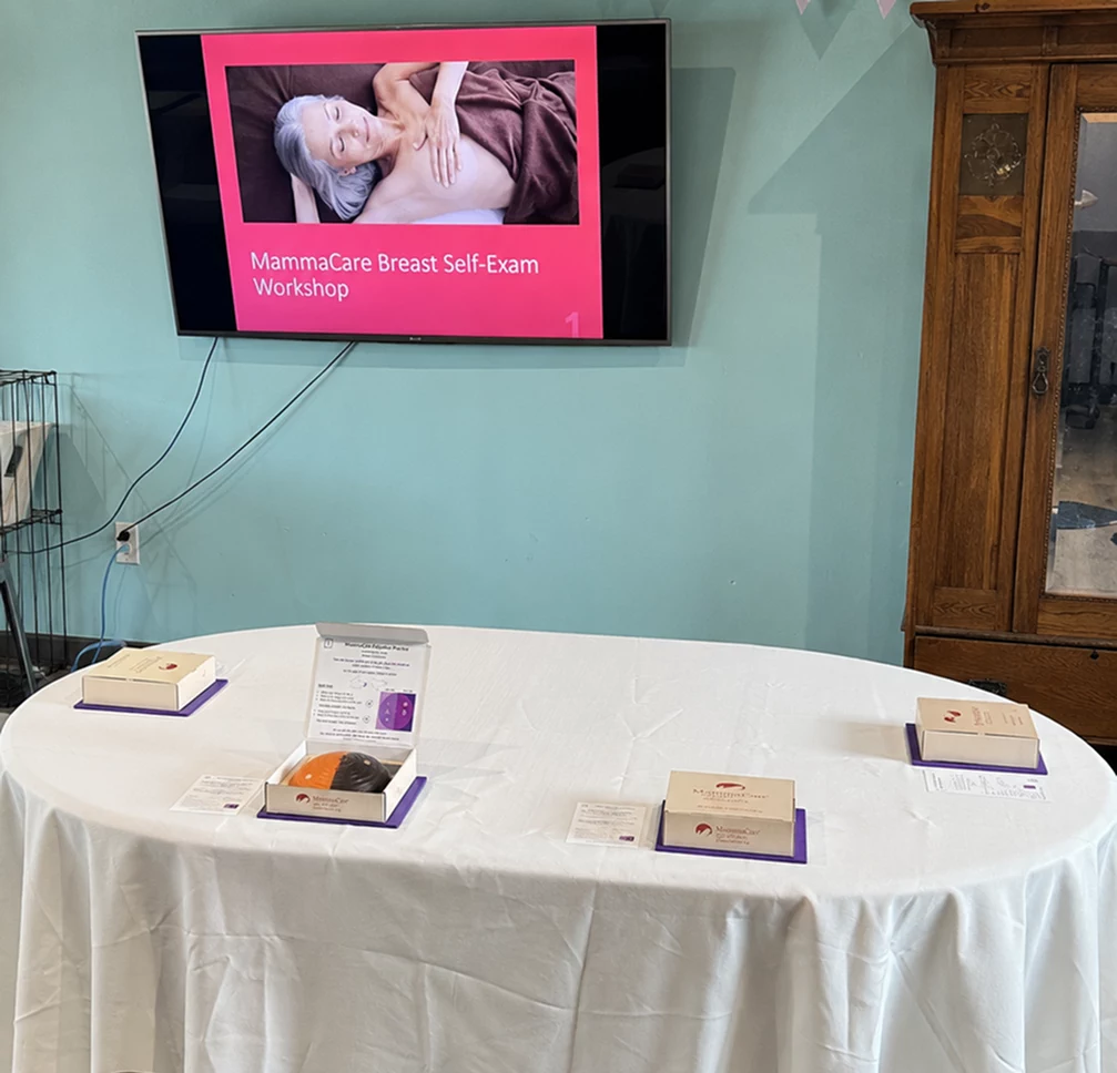 Table set with MammaCare breast models for a breast self-exam workshop, below a screen showing the workshop title slide